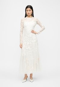 Γυναίκα με λευκό, μακρυμάνικο, μακρύ μέχρι τους αστραγάλους φόρεμα από δαντέλα με λουλούδια, με διαφανή μανίκια και λευκά μυτερά παπούτσια, στέκεται με το χέρι στη μέση.