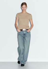 Beige T-shirt met korte mouwen, lichtblauwe brede jeans, bruine riem met zilveren gesp, zwarte haklaarzen, staand tegen een neutrale achtergrond.
