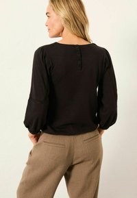 Frau mit blonden Haaren, die eine schwarze Bluse mit langen Ärmeln und Knopfdetails sowie beige strukturierte Hose trägt, mit dem Rücken zur Kamera.