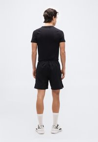 Άνδρας που στέκεται με την πλάτη, φορώντας μαύρο κοντομάνικο πουκάμισο, μαύρα σορτς, λευκές κάλτσες και λευκά αθλητικά παπούτσια σε απλό λευκό φόντο.