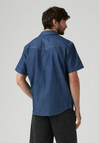 Camisa de mezclilla de manga corta en azul oscuro con textura suave, cuello puntiagudo y dobladillo curvado; presenta detalles de yugo en la parte posterior.