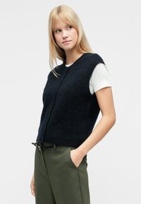 Σκούρο μπλε πλεκτό αμάνικο πουλόβερ με στρογγυλή λαιμόκοψη και κλείσιμο με κουμπιά μπροστά, φορεμένο πάνω από λευκή φανέλα και συνδυασμένο με πράσινο παντελόνι.