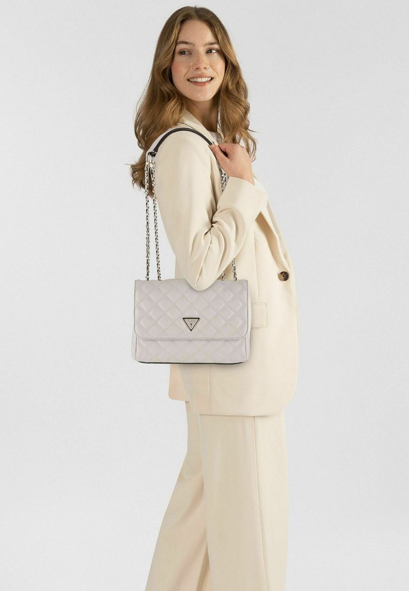 Bolso acolchado gris claro con cierre de solapa, correa de cadena plateada y un adorno de logotipo triangular, llevado sobre un traje color crema.