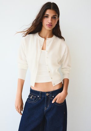 Vrouw met lang donker haar, gekleed in een wit kort vest over een wit topje en donkerblauwe wijde spijkerbroeken, staand tegen een effen achtergrond.