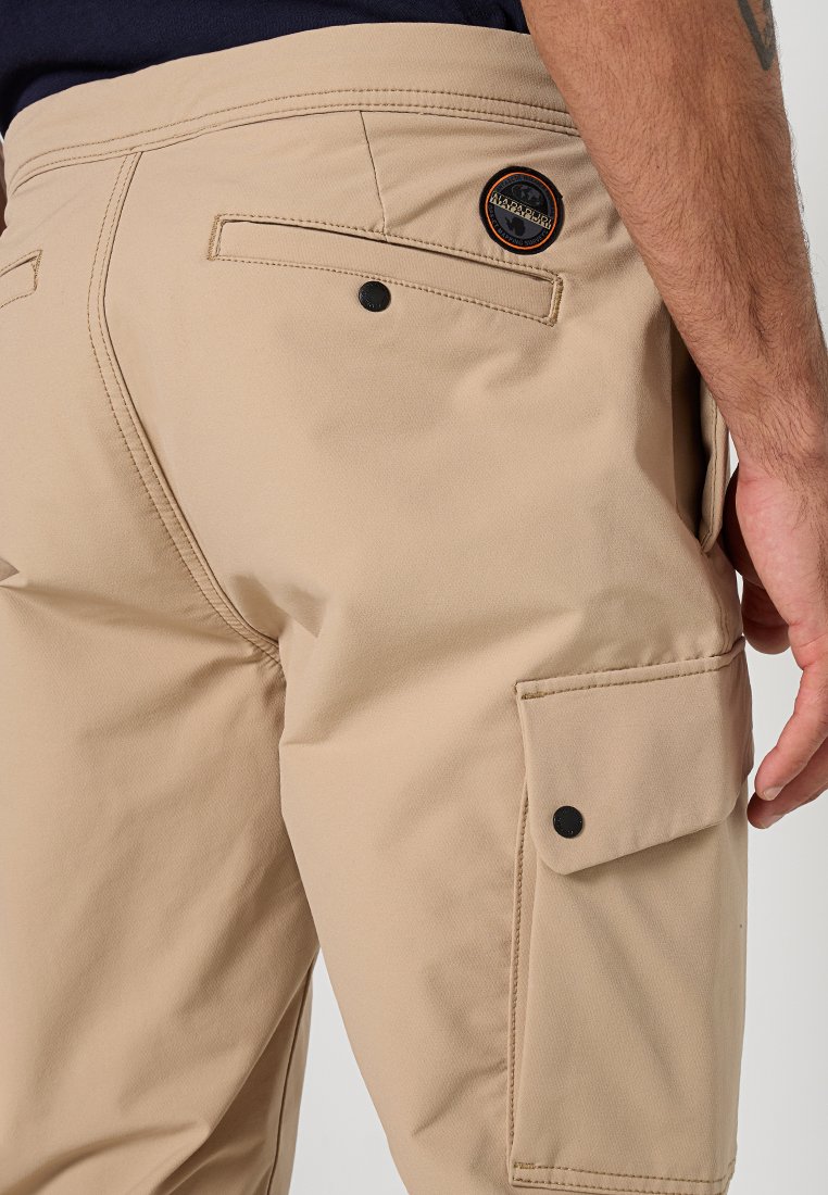 Pantaloni cargo beige con tasca posteriore a pattina e tasca laterale con bottone indossati da una persona con un avambraccio tatuato visibile.