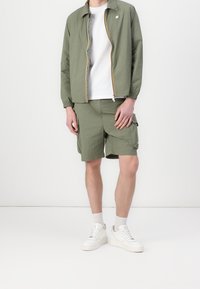 Lightweight olive green zip-up jacket with elastic cuffs, paired with matching cargo shorts. White t-shirt underneath and white sneakers.