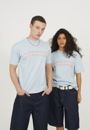 Light blue cotton t-shirts with "LEGACY STUDIOS" text in pink, worn with dark denim shorts. One model has a silver chain necklace.