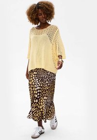Jasnożółty, luźno dziergany sweter z krótkim rękawem, w zestawieniu z długą spódnicą w brązowo-złote wzory; zestaw dopełniają białe buty sportowe.