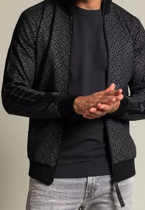 Homme portant une veste noire à motifs zippée sur un t-shirt noir et un jean gris clair, mains jointes à la taille.