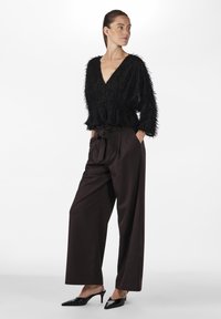 Maglione nero con texture e scollo a V, abbinato a pantaloni marroni a vita alta con una cintura annodata. Tacchi neri a punta e affilati aggiungono contrasto.
