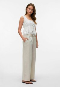 Witte top met kantafwerking en knoopsluiting, gecombineerd met lichtbeige, loszittende broek en zwarte platte sandalen.