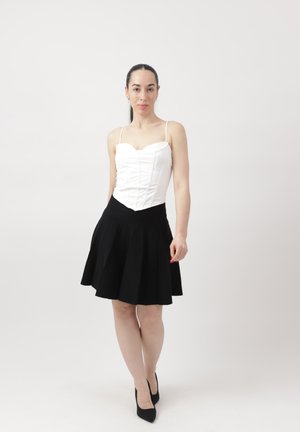 Kobieta stojąca przodem, ubrana w dopasowaną białą bluzkę, czarną rozkloszowaną spódnicę i czarne szpilki na tle jednolitej białej ściany.