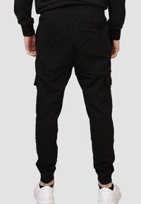 Pantaloni cargo neri con vita regolabile, vestibilità affusolata, tasche laterali e cuciture rinforzate. Indossati con scarpe da ginnastica bianche.