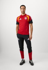 Sarkans un melns Adidas sporta apģērba komplekts ar logotipu, trim svītrām un Vācijas karoga akcentiem uz krekla un biksēm. Melnas kurpes.