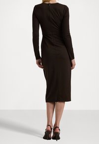 Hnědá midi šaty s dlouhým rukávem a přiléhavým střihem, se zipem na zádech a bočním rozparkem, doplněné vysokými otevřenými botami.