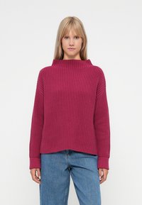 Sweater malha magenta com colarinho alto e largo e textura canelada; apresenta ombros caídos e mangas longas. Usado com jeans azuis.