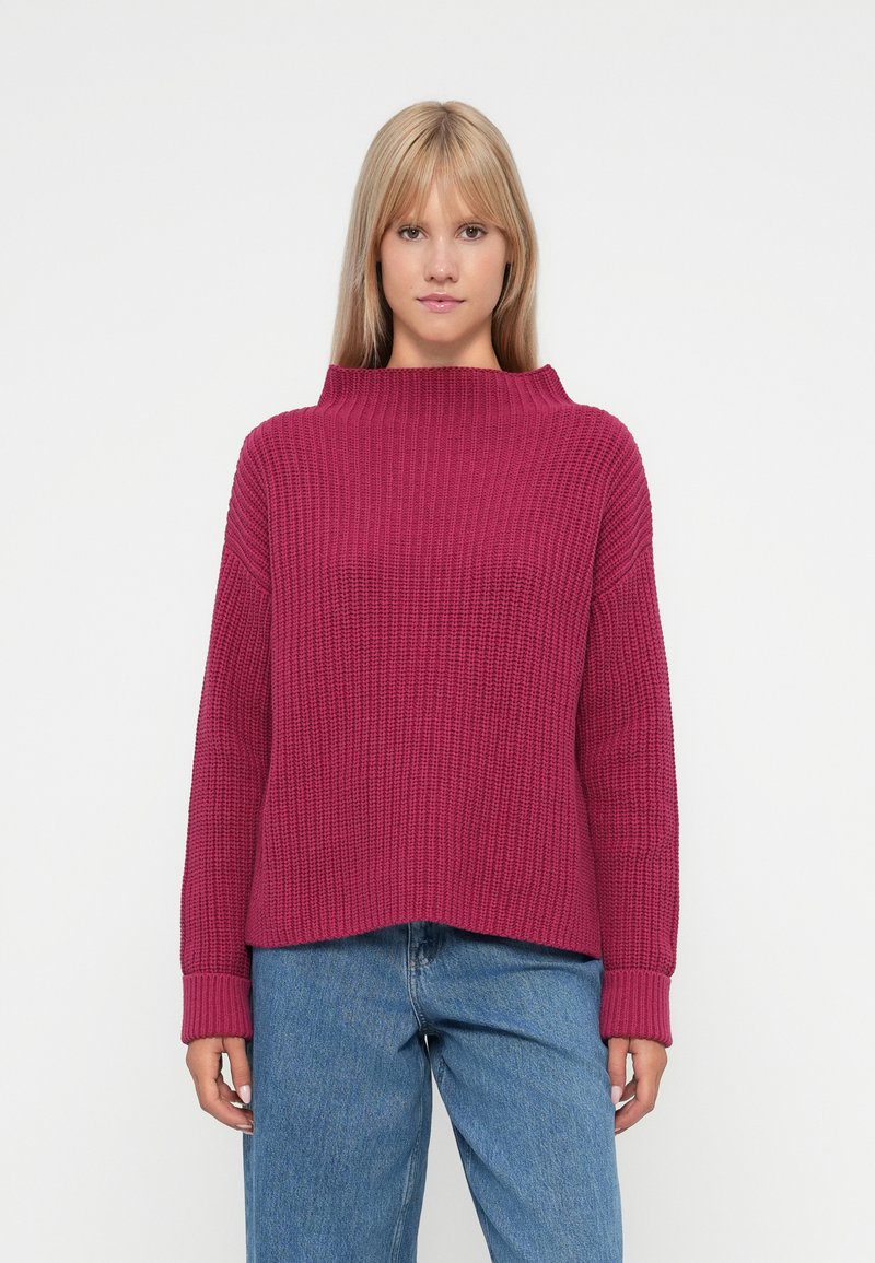 Sweater malha magenta com colarinho alto e largo e textura canelada; apresenta ombros caídos e mangas longas. Usado com jeans azuis.