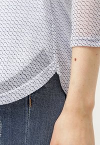 Valkoinen läpikuultava paita, jossa on pieniä laivastonsinisiä kuvioita, pyöristetty helma ja kolmannen neljänneksen hihat, yhdistettynä sinisiin farkkuihin.