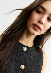 Schwarzes, strukturiertes Kleid mit drei auffälligen goldenen Knöpfen an der Vorderseite, das ein tailliertes Design und einen minimalistischen Ausschnitt präsentiert.