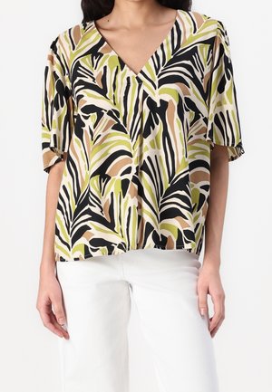 Kobieta ubrana w luźną bluzkę z krótkim rękawem i dekoltem w serek z abstrakcyjnymi liściastymi wzorami w kolorach czarnym, oliwkowozielonym, beżowym i białym, w połączeniu z białymi spodniami.