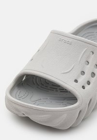 Grå gummiskidsandal med en texturerad ovandel, öppen design, ventilationshål och en formad fotbädd. Varumärkeslogotyp subtilt präglad på remmen.