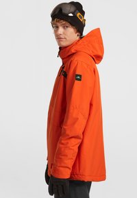 Felkleurige waterdichte jas met een capuchon, zwarte accenten en merk op de mouw. Draagt donkere handschoenen en heeft een bril op het voorhoofd.