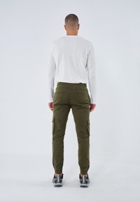 Zelené cargo kalhoty s elastickým pasem a bočními kapsami, s dlouhým rukávem bílé tričko. Model je vidět z pohledu zezadu, má na sobě šedé boty.