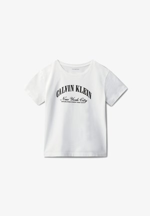 Enfant aux cheveux bouclés portant un t-shirt blanc Calvin Klein, une jupe en jean et des mocassins noirs, debout les mains dans les poches.