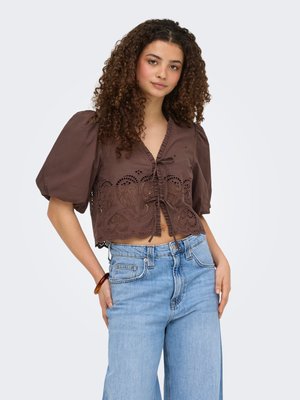 Femme aux cheveux bouclés portant un top court en dentelle à œillets marron et un jean taille haute bleu clair, posant avec les mains dans les poches.