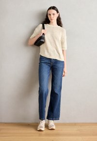 Krémový pletený svetr s krátkým rukávem, nošený s modrými kalhotami s širokými nohavicemi. Modelka drží černou kabelku; béžové tenisky doplňují outfit.