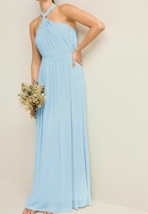 Kvinde iført en lang, lyseblå halterneck-kjole, der holder en buket tørrede blomster, står mod en ensfarvet beige væg.