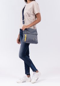 Borsa a tracolla grigia conpatta a ribalta, accentuato da una cerniera gialla e un tessuto testurizzato. Indossata con jeans blu e sneaker bianche.