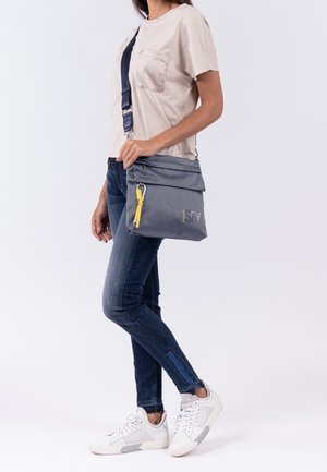Sac messager gris avec un rabat repliable, accent de zip jaune et tissu texturé. Porté avec un jean bleu et des sneakers blanches.