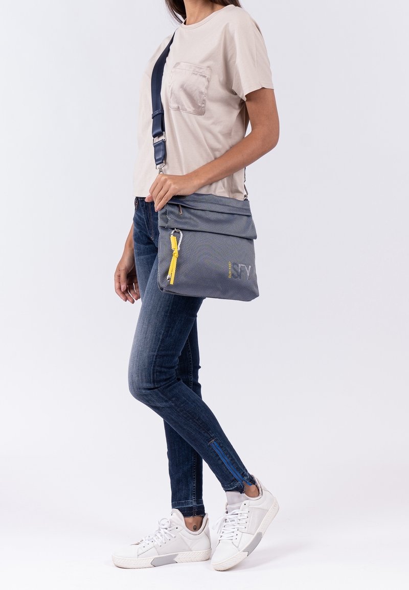 Borsa a tracolla grigia conpatta a ribalta, accentuato da una cerniera gialla e un tessuto testurizzato. Indossata con jeans blu e sneaker bianche.