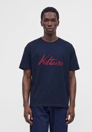 Jeune homme portant un t-shirt bleu marine avec un texte rouge en cursive "Voltaire" et un jean bleu foncé, debout devant un fond clair uni.