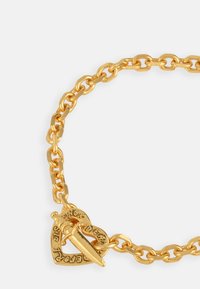 Gold chain bracelet featuring oval links and a heart-shaped charm with a dagger accent. The charm is engraved with decorative text.