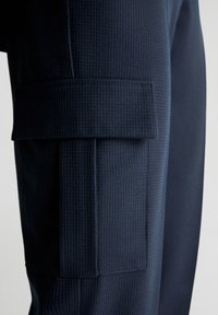 Morskie spodnie cargo z teksturowanej tkaniny posiadają dużą boczną kieszeń z klapką oraz detalami zszycia, podkreślając elegancki, dopasowany krój.
