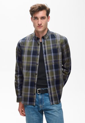 Ung mand med kort brunt hår iført en grøn og marineblå ternet skjorte med knapper over en sort t-shirt og blå jeans med sort bælte.