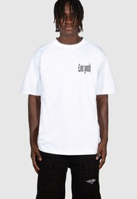 Vitt bomulls t-shirt med svart text som läser "Lost youth" på vänster bröst. Lös passform, rundad hals, kortärmad, buren med svarta cargo-byxor.