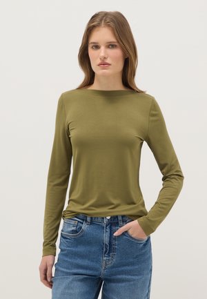 Frau mit hellbraunen Haaren, die ein olivgrünes langärmliges Oberteil und blaue Jeans trägt, mit einer Hand in der Tasche, vor einem einfarbigen Hintergrund stehend.