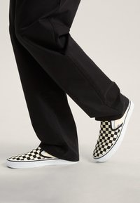 Sorte og hvide ternede slip-on sneakers med overdel i kanvas og hvid gummisål, parret med sorte bukser i sidevisning.