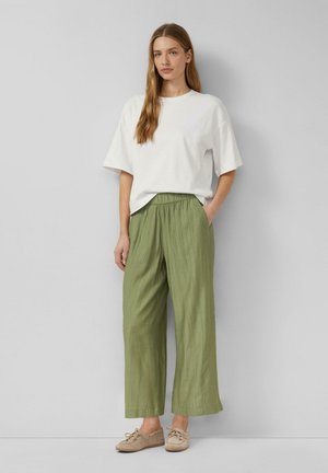 Kobieta stojąca, ubrana w luźną białą koszulę z krótkim rękawem, oliwkowo-zielone szerokie spodnie i beżowe buty sznurowane na tle szarego tła.