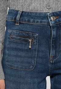 Jeans in denim blu, con una tasca con cerniera, caratterizzati da cuciture a contrasto e vita aderente. Il tessuto appare testurizzato e leggermente usurato.