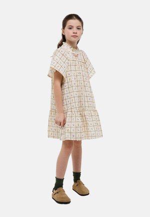Jeune fille debout portant une robe ample crème à manches courtes avec un motif floral en grille, des chaussettes vert foncé et des chaussures beiges à enfiler.