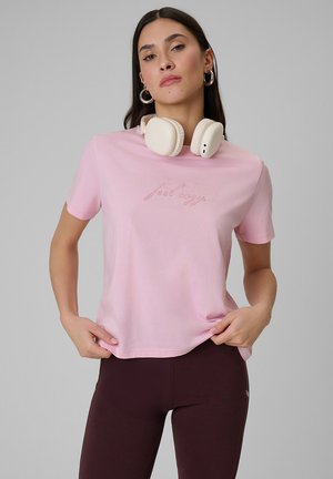 Młoda kobieta o długich ciemnych włosach, ubrana w różowy t-shirt i ciemne legginsy, trzyma brzeg koszulki, a na szyi ma beżowe słuchawki.