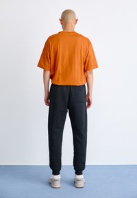 Mand med barberet hoved står overfor en hvid væg iført en orange t-shirt, sorte træningsbukser og hvide sneakers på et blåt gulv.