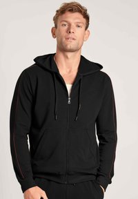 Zwarte rits-hoodie met zijzakken, voorzien van een rode accentstrip langs de mouwen en een capuchon met trekkoord. Zachte stof, ontspannen pasvorm.