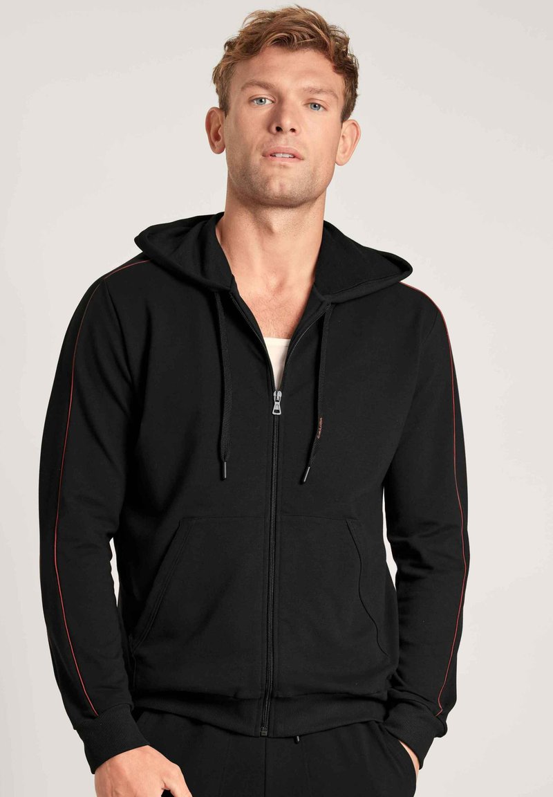 Zwarte rits-hoodie met zijzakken, voorzien van een rode accentstrip langs de mouwen en een capuchon met trekkoord. Zachte stof, ontspannen pasvorm.