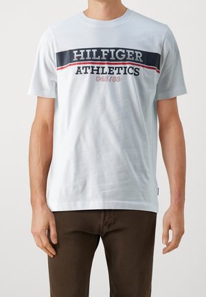 Mężczyzna w białej koszulce z krótkim rękawem z napisem „HILFIGER ATHLETICS 045/88” w granatowym i czerwonym kolorze, w połączeniu z ciemnobrązowymi spodniami.