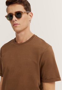 Braunes T-Shirt mit kurzen Ärmeln und Rundhalsausschnitt sowie geprägtem Logo. Das Modell trägt eine Sonnenbrille mit transparentem Rahmen und dunklen Gläsern.